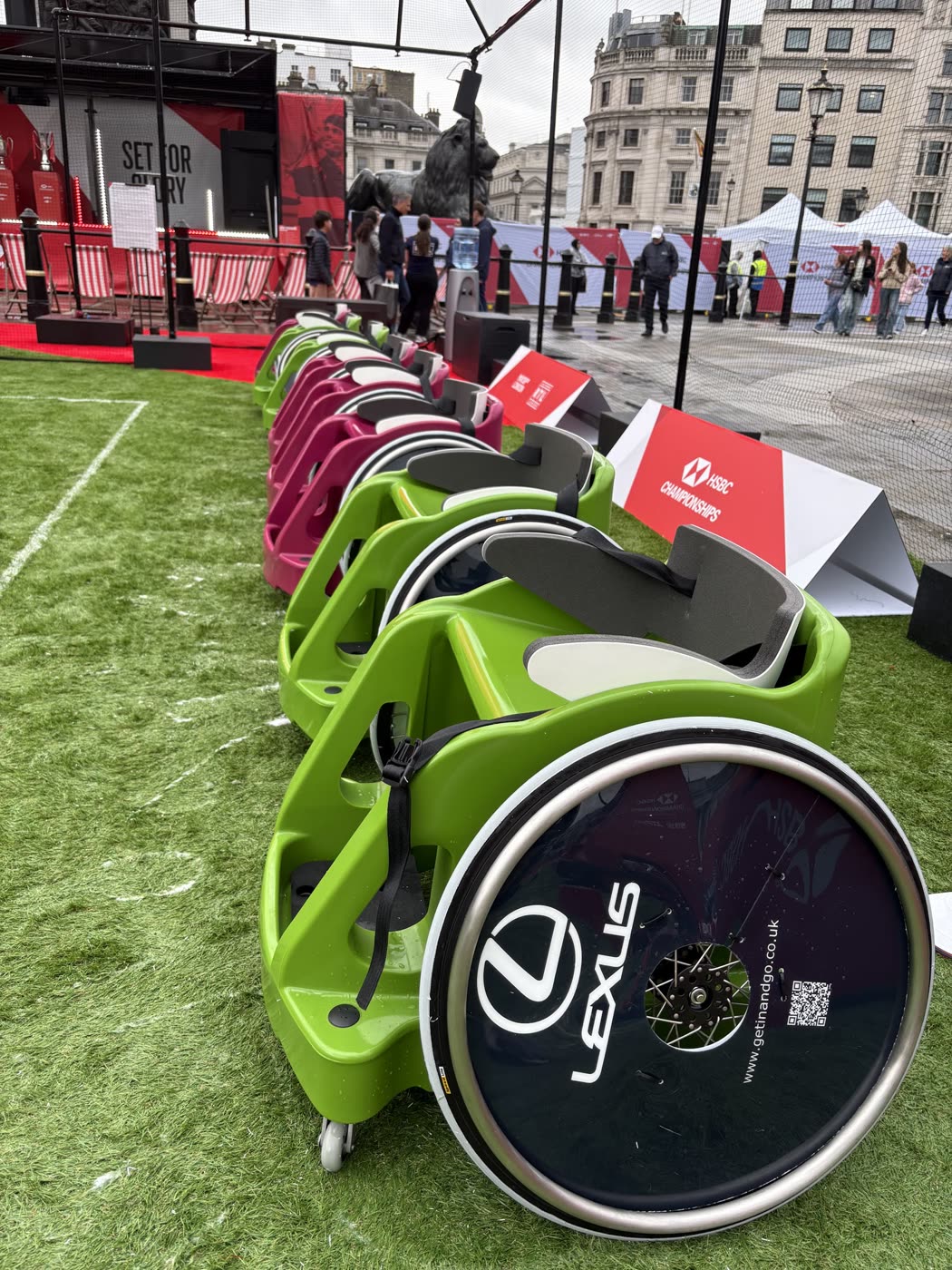 A row of green and pink PlayMoovin' sport wheelchairs lined up on the grass court at Trafalgar Square