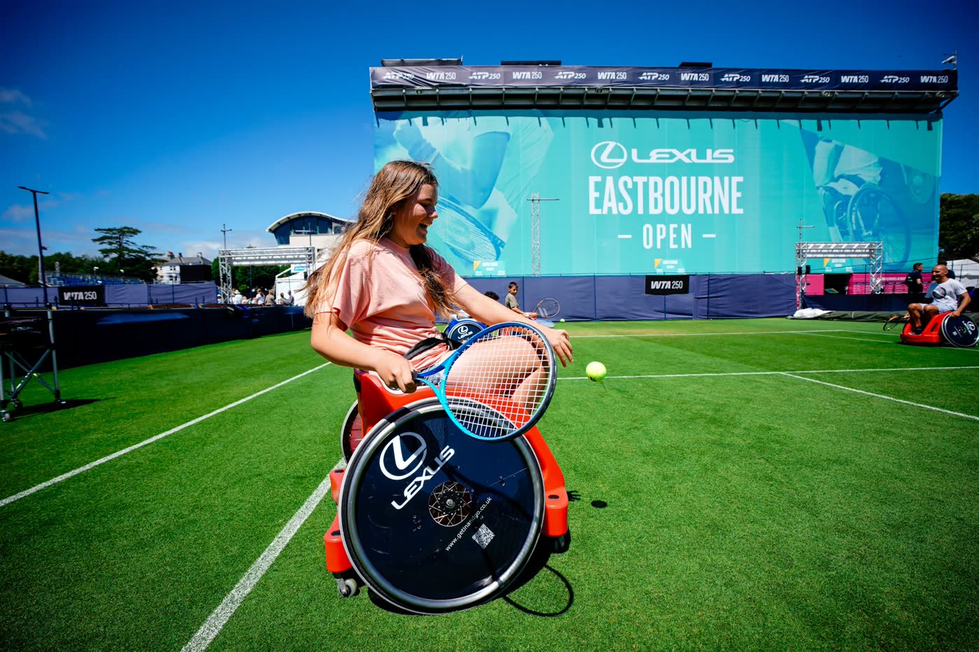 A girl swings a racket at the ball from a red sport wheelchair on a grass court, in front of a large Lexus Eastbourne Open banner.