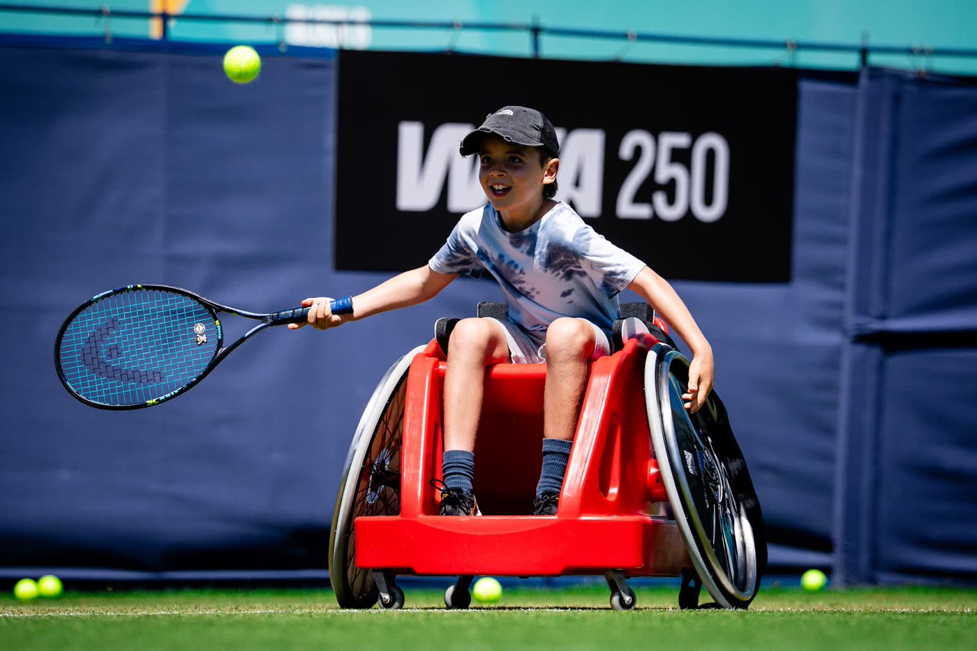A boy in a red sport wheelchair stretches out to play a forehand, eyes fixed on the ball, in front of a WTA 250 board.