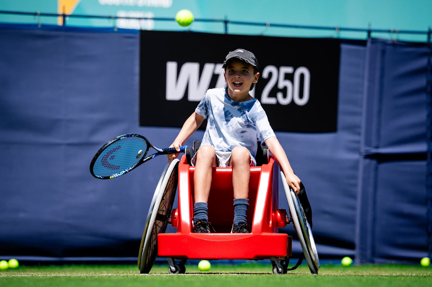 A boy grins as he lines up a shot in a red sport wheelchair, tennis balls scattered across the grass around him.