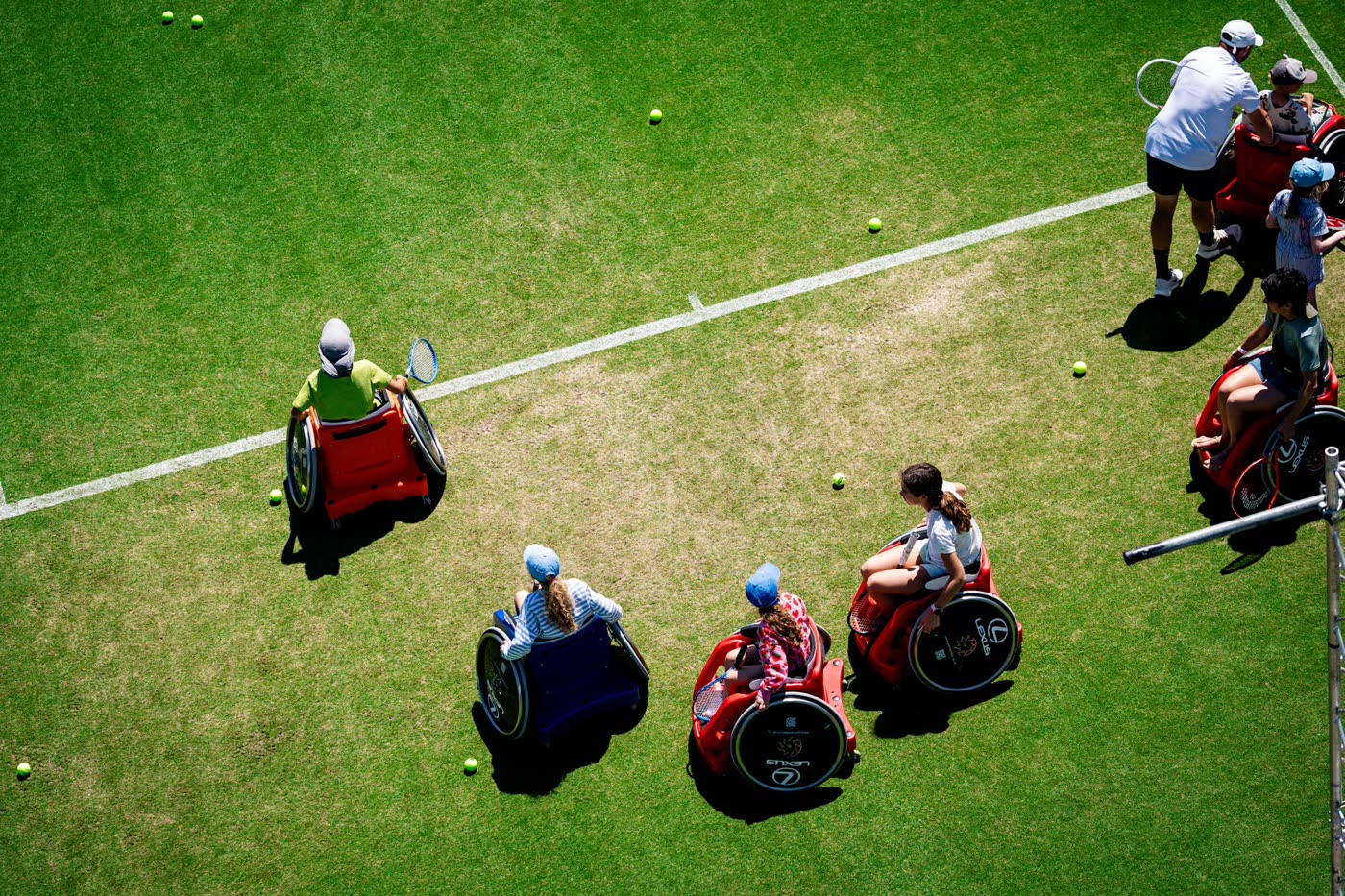 Overhead view of five children spread across a sunlit grass court in red, blue and orange sport wheelchairs, a coach standing alongside them.