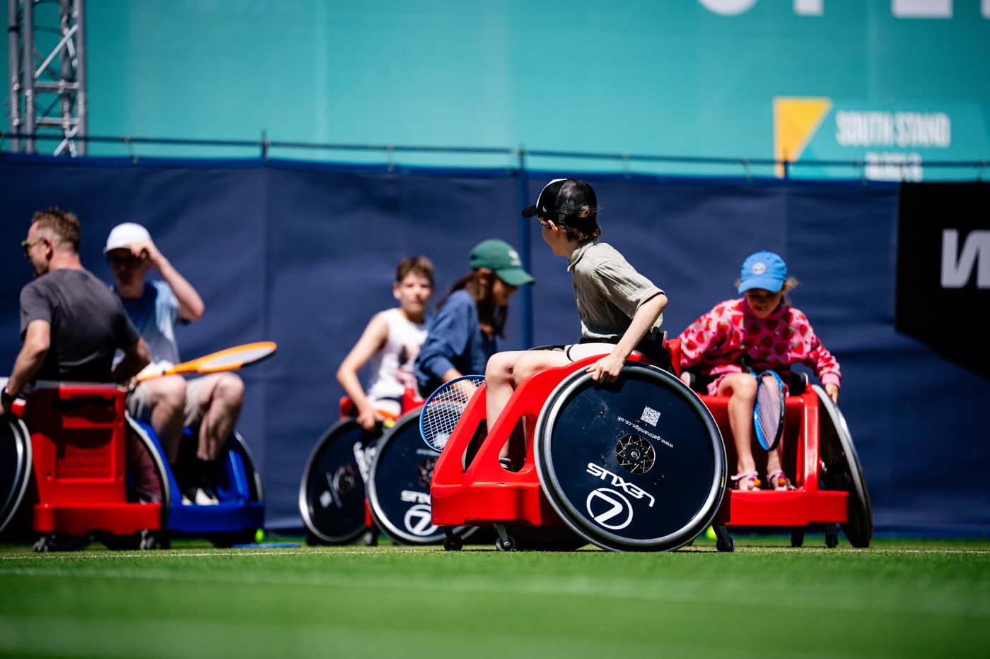 Children and adults courtside in red and blue sport wheelchairs, rackets resting across their laps